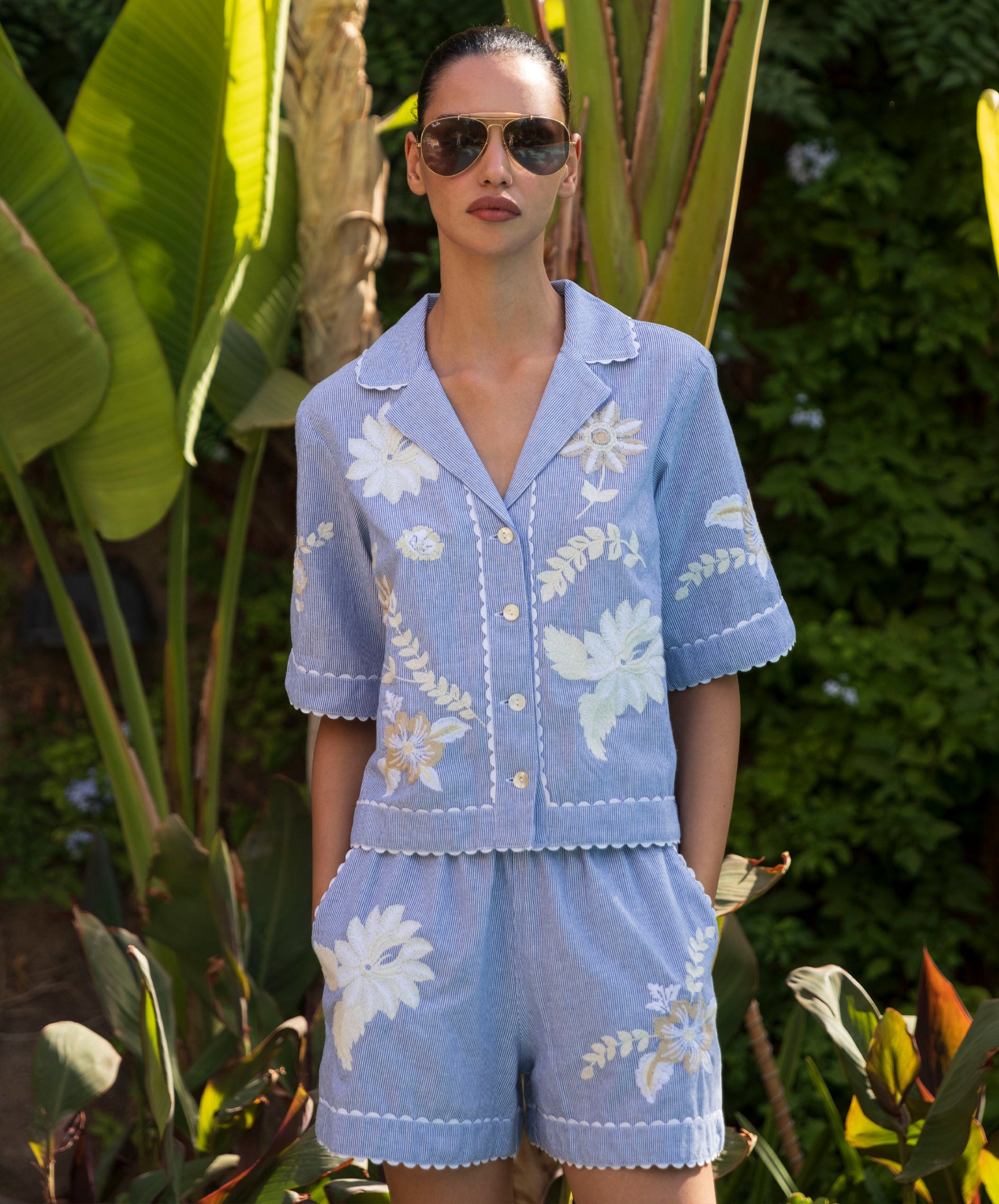 Woman wearing a blue striped embroidered blouse and shorts set with sunglasses in a garden setting