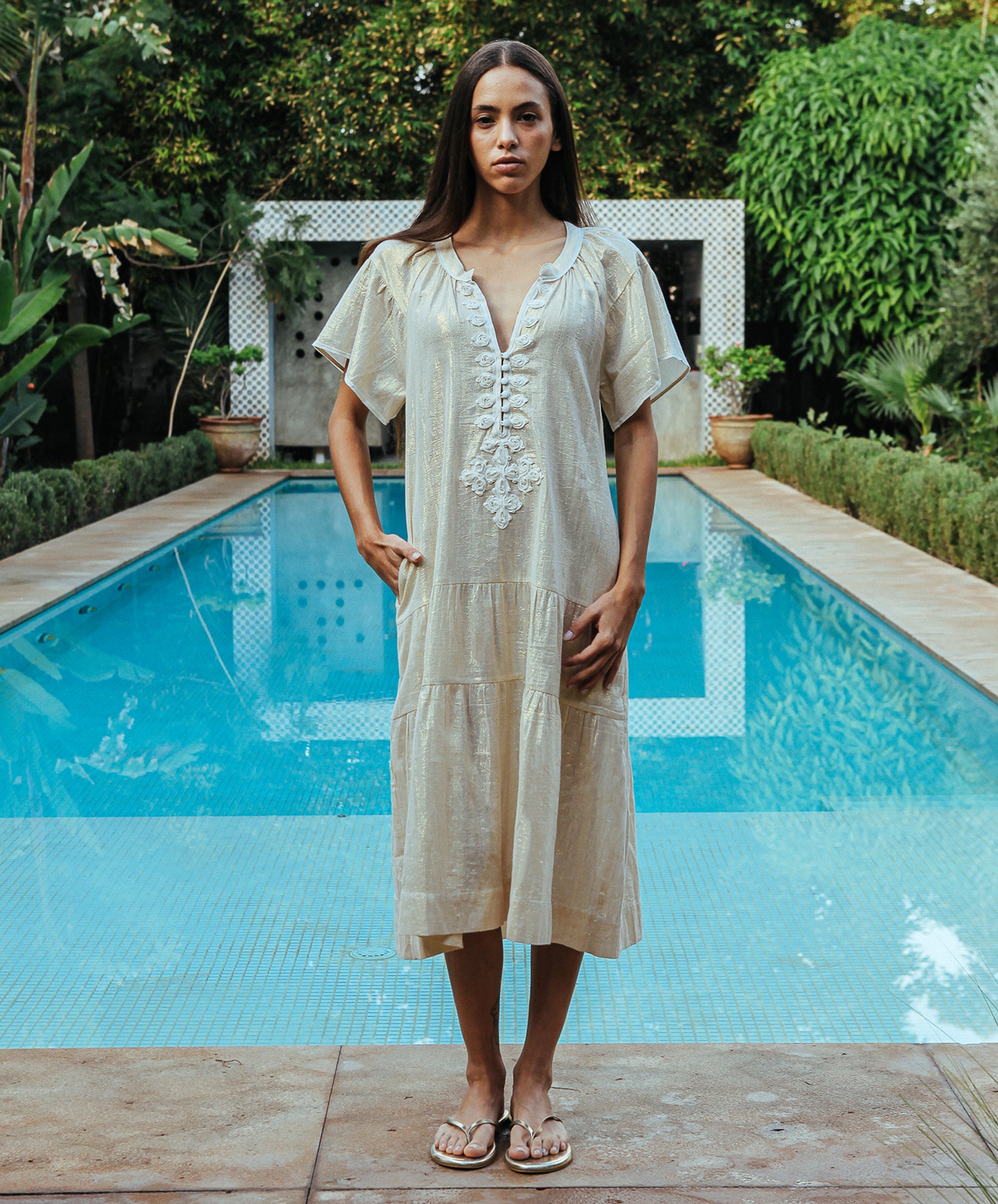 A model stood in front of a swimming pool wearing a Rose and Rose Uxbridge gold dress with white passementerie decoration. 