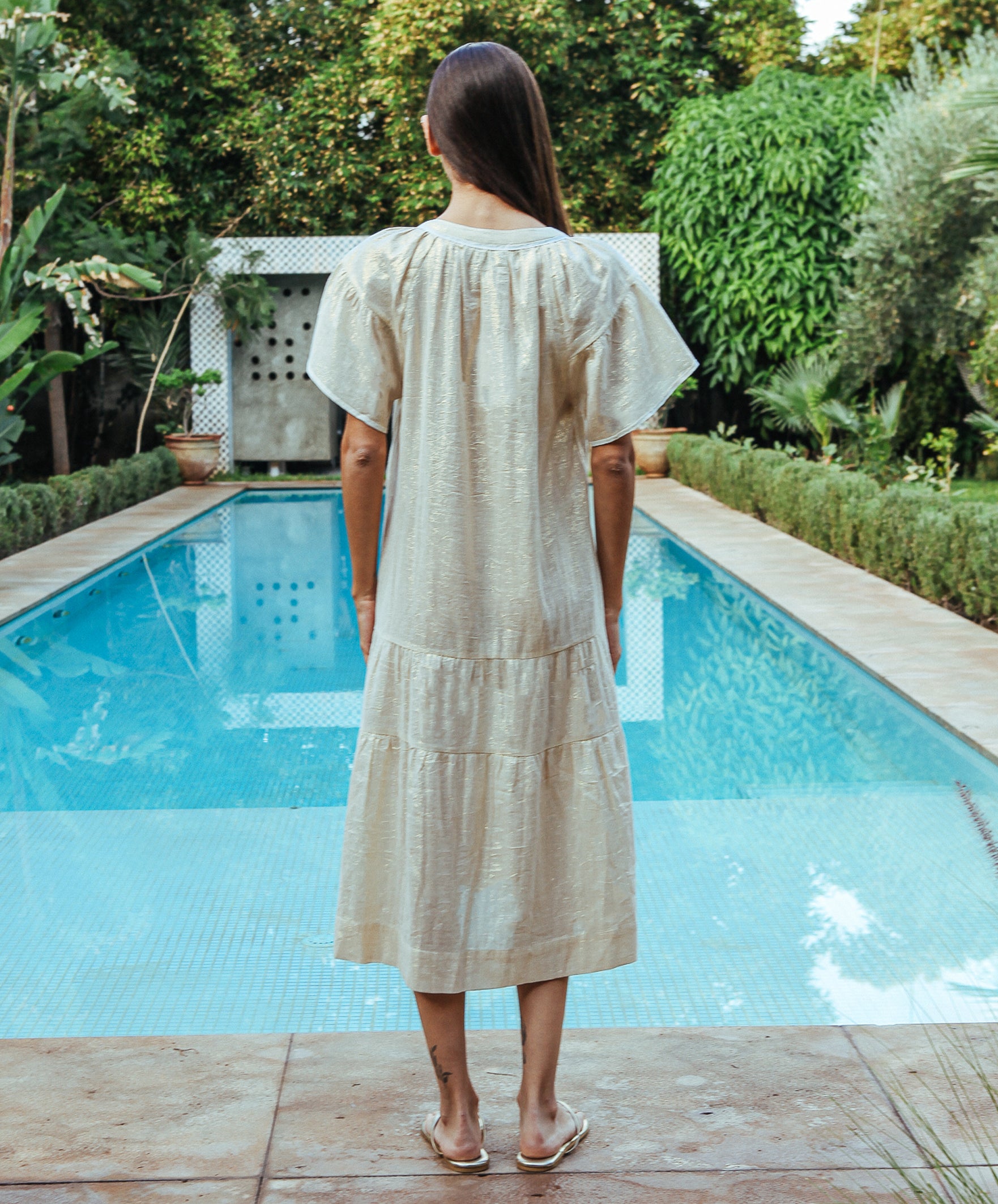 A back view of a model stood in front of a swimming pool wearing a Rose and Rose Uxbridge gold dress. 