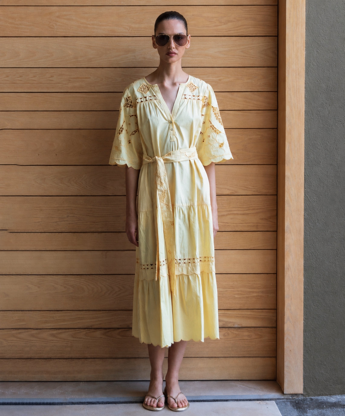 Woman wearing a light yellow embroidered dress and straw hat against a wooden wall.