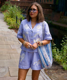 Influencer @irenadworld wearing a light blue striped short-sleeve button-up shirt and matching shorts set with white floral embroidery, styled with a blue and white tote bag, sunglasses, and bracelets, standing outdoors on a stone path.