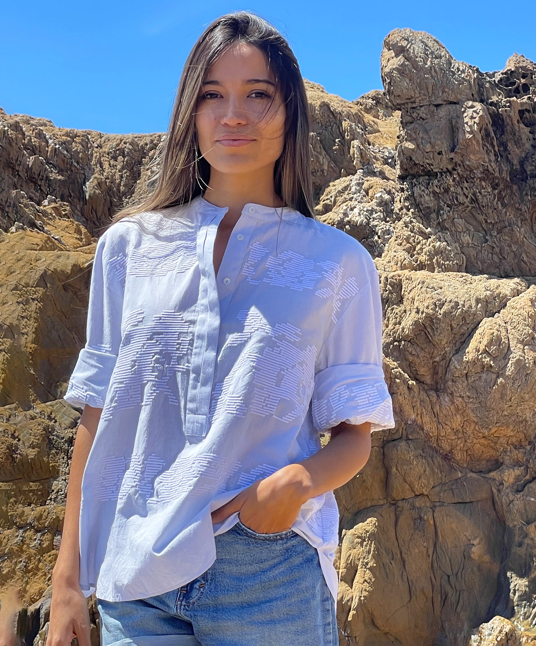 A model stood in front of rocks wearing a pale blue embroidered Parma top with denim shorts. 