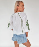 Back view of a beautiful long blond haired model wearing a premium white cotton top with olive green appliqué motifs, and denim shorts.